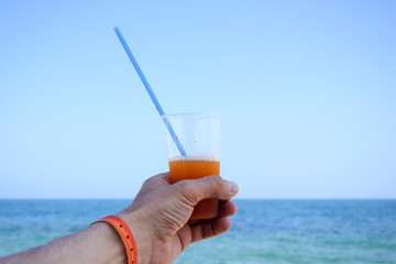 Glass with an orange cocktail on background of the sea. Male hand holds glass with plastic tube.