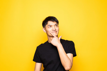 Young asian chinese manwith hand on chin thinking about question, pensive expression standing over isolated yellow background . Smiling with thoughtful face. Doubt concept.