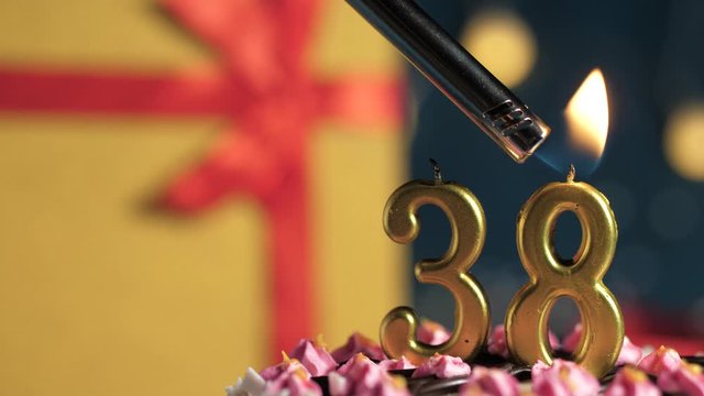 Birthday cake number 38 golden candles burning by lighter, blue background gift yellow box tied up with red ribbon. Close-up and slow motion