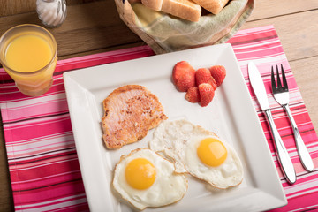 American Breakfast sunny side up eggs bacon toasted bread fruits and juice on a simple colourful mantel on wooden table