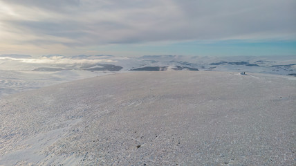 A aerial scenic view of a snowy mountain summit with mountain range in the background under a majestic blue sky and some altitude white cloud