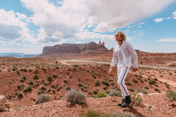 A curly haired blonde man posing around the famous Buttes of Monument Valley from Arizona, USA, wearing white linen shirt and white pants, black shoes, majestic beauty.