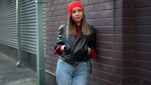 Beautiful European girl dressed black leather jacket and jeans and red hat standing backyard Hands in pockets leaning against red brick wall and smiling youth culture modern woman 30s year autumn 
