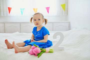 Adorable toddler girl in blue dress sitting on bed with pink hyacinths and white wooden number two