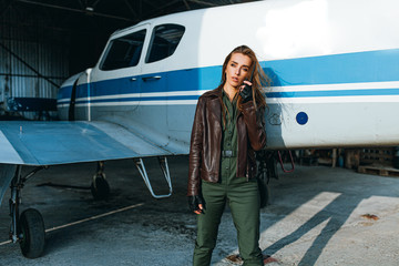 stylish girl in the guise of a pilot talking on the walkie-talkie in the hangar garage against the background of the plane in a leather jacket, green overalls and gloves