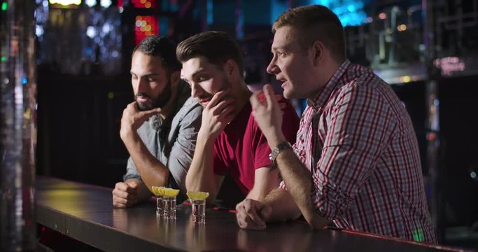 Three young men standing at bar counter and talking with bartender. Barkeeper giving short drinks to Middle Eastern and Caucasian fellows. Male friendship, resting, lifestyle. Cinema 4k ProRes HQ.