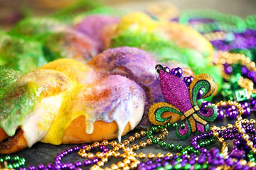 Mardi Gras king cake with yellow, green, and purple sprinkles surrounded by Mardi Gras beads and a glittering fleur de lis.
