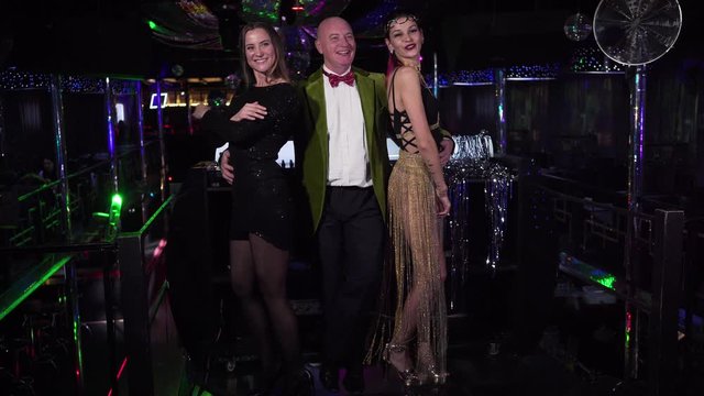 Elderly Caucasian man and two young women posing in night club. Cheerful group of people smiling and talking in disco. in front of DJ controller. Entertainment, lifestyle, club workers.