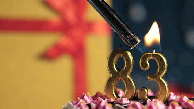 Birthday cake number 83 golden candles burning by lighter, blue background gift yellow box tied up with red ribbon. Close-up and slow motion