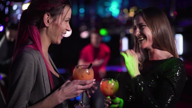Close-up of two young pretty Caucasian girls looking back at visitors in night club, talking and laughing. Joyful women spending evening with cocktails in disco. Leisure, lifestyle, joy.