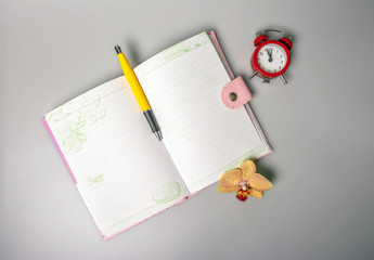 An open notebook, pen, orchid flower and an alarm clock reminiscent of time are on the table. Office. School supplies. Place for text. View from above.