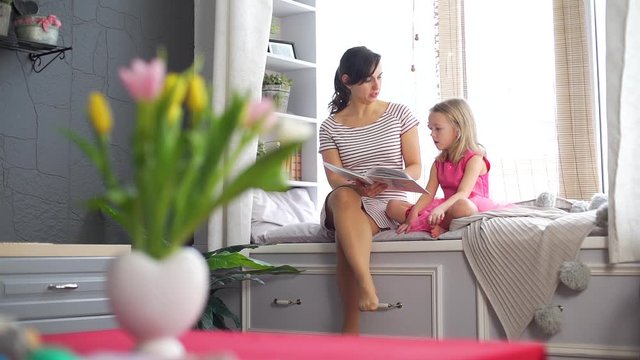 Young Mother Reading Book to her Cute Little Daughter at Home. They Sitting on the Big Cozy Windowsill. People, Lifestyle and Happy Family Concept