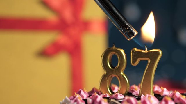 Birthday cake number 87 golden candles burning by lighter, blue background gift yellow box tied up with red ribbon. Close-up and slow motion