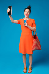 joyful girl in a bright dress takes a selfie, holds a cocktail and bag in her hands, the concept of leisure and travel