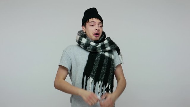 Flu-sick man in hat and scarf sneezing in napkin, cleaning his running nose and crying with funny comical expression, feeling unwell suffering influenza. indoor studio shot isolated on gray background