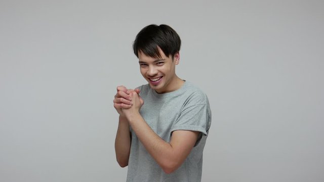 Young cunning smart guy in T-shirt rubbing hands and smiling slyly, thinking devious evil idea, genius plan to prank friend, smirking and conspiring. indoor studio shot isolated on gray background