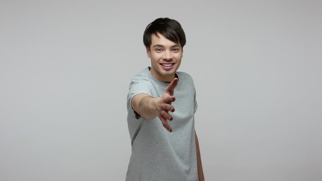 Positive man in T-shirt holding out palm to camera, giving hand to handshake and smiling amiably, young applicant greeting at job interview, getting acquainted. indoor studio shot gray background.