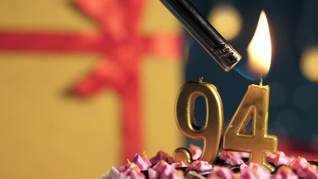 Birthday cake number 94 golden candles burning by lighter, blue background gift yellow box tied up with red ribbon. Close-up and slow motion