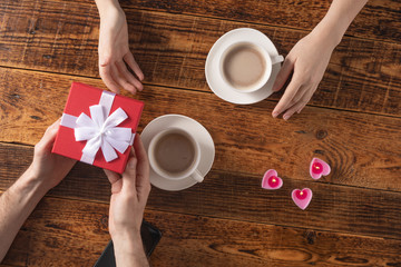 Valentine's Day celebration concept. A nice gift for your loved one. Hands of man and woman with coffee mugs on a wooden table background. Copy space. Flat lay. Close-up.