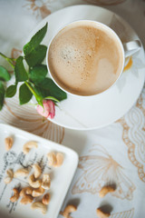 Top view of a tablecloth covered table with a cup of delicious morning coffee and a plate of cashew nuts. Garden rose for mood. Toning