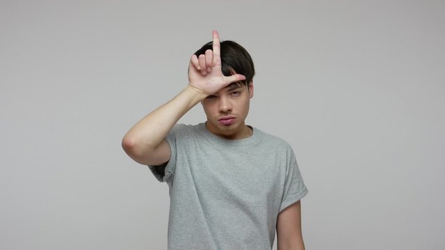 Unhappy gloomy guy in T-shirt showing loser gesture with L sign on forehead, looking desperate and depressed about failure, unemployment, fired from job. indoor studio shot isolated on gray background