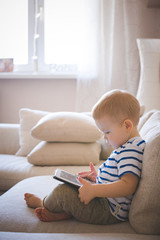 A little cute boy sits on a sofa in a living room at home and works on a tablet. The child plays educational games and programs. Early development. Toning and artificial noise