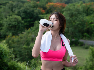 Close up Young asian woman drink water after running on road in the morning sunshine outdoors. Female jogger exercising. Fitness and healthy lifestyle