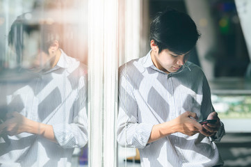 Portrait of young man using smartphone while standing near window glass with sunlight.