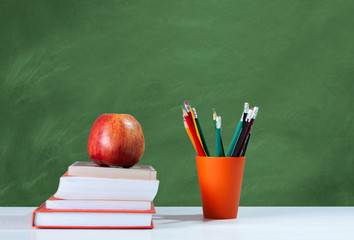 Back to school, orange pencil holder, stack of books on white table with red apple, empty green school board background, education concept.