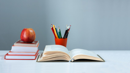 Orange pencil holder, open hardback book, diary, fanned pages on stack of books on white table with red apple. Books stacking. Back to school concept. Copy Space. Education learning background.