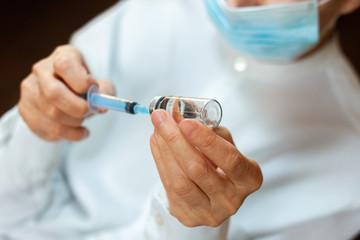 doctor in a white surgical gown and blue mask holds in hands a medical syringe and bottle with...