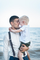 Young father holds child on hands against sea. A kid hugs dad. Vertical frame