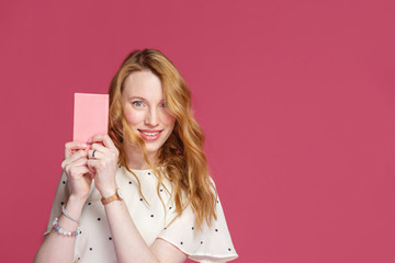 Cheerful blonde girl holds a pink notebook in her hands, looks at the camera and smiles, isolated on a pink background with copy space.