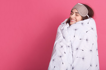 Horizontal portrait of cheerful sleepy attractive brunette closing eyes, having pleasant smile, wearing sleeping mask, having blanket round body, being in high spirits. Copyspace for advertisement.