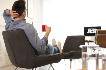 Young man with cup of drink relaxing in comfortable chair at workplace