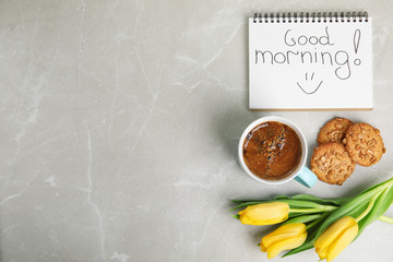 Delicious coffee, cookies, flowers and GOOD MORNING wish on grey table, flat lay. Space for text