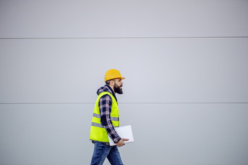 Side view of hardworking construction worker with safety helmet on head and in vest holding laptop...