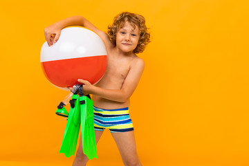 Little boy with curly hair in swimsuit with fins and big rubber ball isolated on yellow background