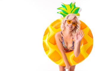 Pretty blonde caucasian female stands in swimsuit with rubber beach pineapple ring and smiles isolated on white background