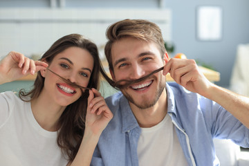 Portrait of cute young playful couple teasing with fake mustache sitting in sofa.
