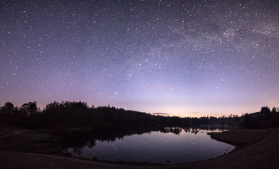 Milky Way over a lake with stars reflecting in the water with a purple sky and surrounded by trees