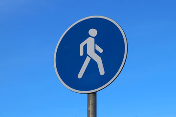 Prescriptive road sign in blue with a round shape with a white person walking, indicating a footpath against a blue sky. Selective focus.