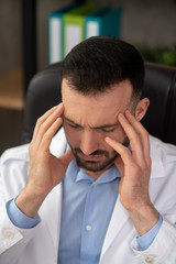 Dark-haired bearded doctor in a white robe having a headache