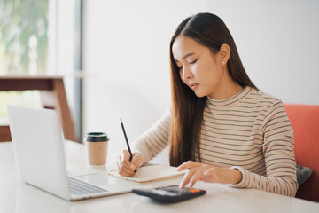 Young Asian woman using a calculator and other hand using pencil take a note in the book and have notebook on the table at cafe or co working space. co working space concept.