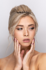 Fototapeta premium A large head portrait of a beautiful young girl with dyed blonde hair arranged in a hairstyle, with makeup and beautiful lips and teeth. Hands near the face. a neutral gray background