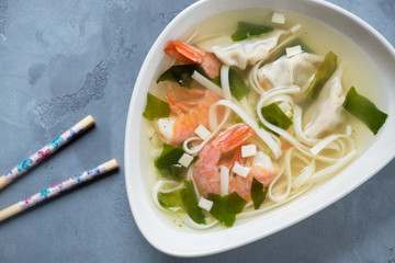 Bowl of wonton soup with noodles and shrimps on a grey concrete surface, view from above, close-up
