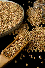 Scattered green buckwheat groats with wooden scoop , grey plate and glass jar on black background. Vertical photo