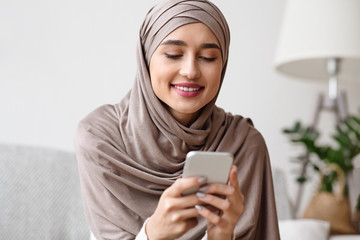 Modern Muslim Girl In Hijab Typing Message On Smartphone At Home