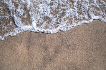White foamy wave hitting the sandy beach shore on a hot summer day.
