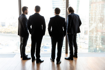 Four confident businessmen back in suit standing looking at window.